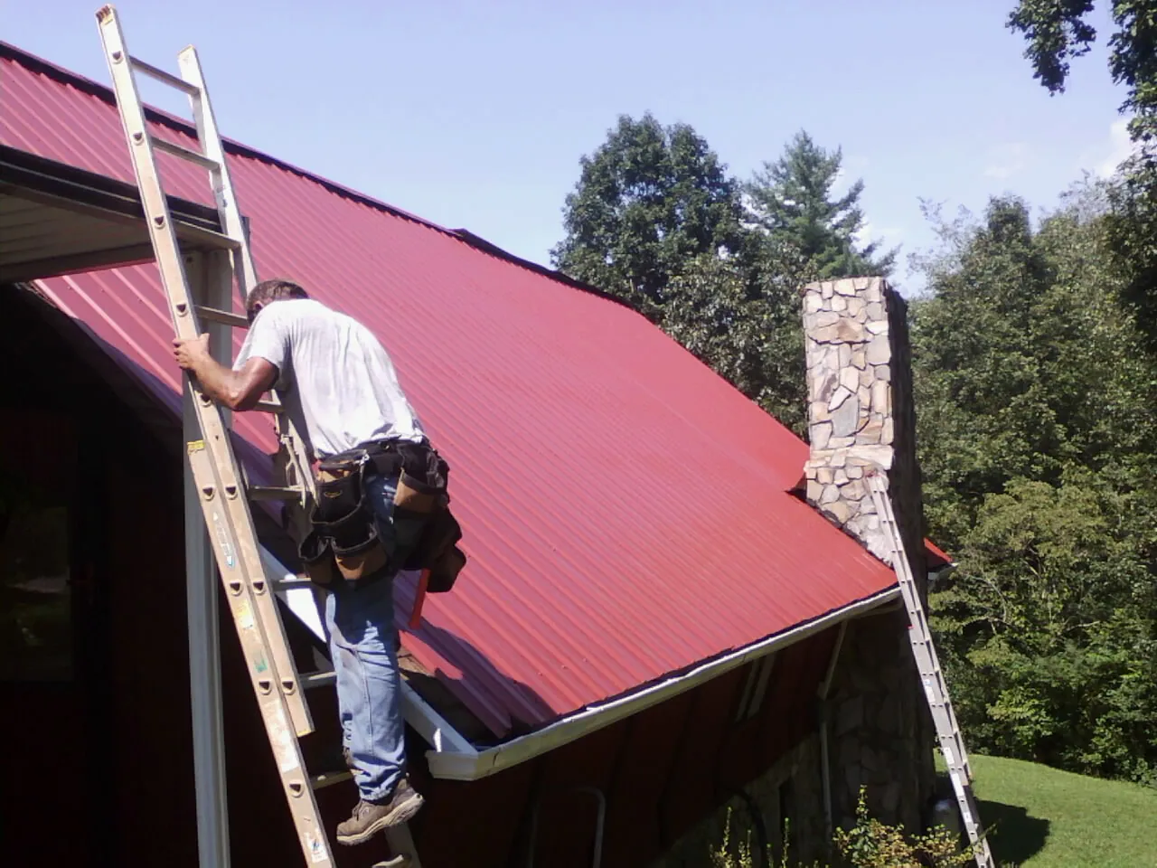 Roofer on a ladder beside a red metal roof and stone chimney — Soffit & Fascia Repair by Roofing Brothers in Oneonta